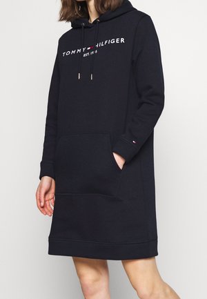 Kobieta ubrana w czarną sukienkę z kapturem Tommy Hilfiger z przednią kieszenią i logo na piersi, stoi z jedną ręką w kieszeni.