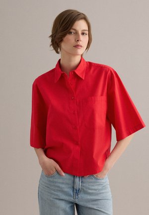 Giovane con capelli corti castani che indossa una camicia rossa brillante a maniche corte abbottonata e jeans azzurri chiari, in piedi con le mani nelle tasche.