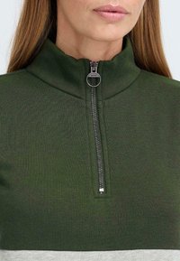 Groene rits-top met hoge kraag, geribbelde structuur en een horizontale grijze streep aan de onderkant; zilveren ritsdetail zichtbaar.