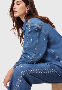 Chaqueta de mezclilla y pantalones con remaches plateados a lo largo de las costuras, en un color azul medio. La tela texturizada y el corte relajado son prominentes.