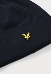 Marineblå ull lue med brettet oppkant, som har en gul brodert ørn logo foran. Myk tekstur og enkelt design.