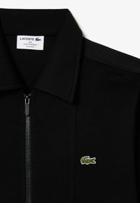 Chaqueta negra con cremallera y cuello, que presenta un logo de cocodrilo bordado en verde, confeccionada en una tela suave con costuras visibles.