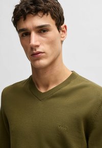 Maglione verde a maglia con scollo a V e una texture sottile; presenta il logo ricamato "BOSS" sul petto. Silhouette morbida e aderente.