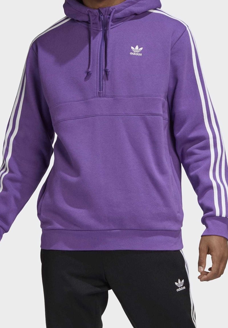 Lila hoodie med vita ränder längs axlarna, med dragkedja i halsen, logotyp på bröstet, tillverkad av mjukt tyg, kombinerad med svarta byxor med logotyp.