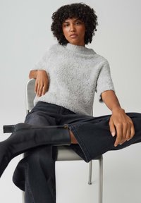 Maglione grigio chiaro, lavorato a maglia, con maniche corte abbinato a jeans scuri a gamba larga e stivali neri fino al ginocchio. Seduta su una sedia di metallo.