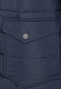 Tmavě modrá vatovaná bunda s velkou náprsní kapsou, která má kovový knoflík s motivem kotvy a hladkou texturu.