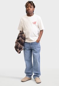 T-shirt beige avec du texte rouge, jeans en denim bleu et chaussures beiges. Tenant une veste en plaid marron et noir, sous une casquette noire.