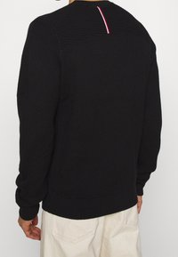 Maglione nero a maglia con parte superiore strutturata e maniche a costine. Presenta un piccolo accento rosso e bianco nella parte posteriore del collo. Vestibilità leggermente ampia.