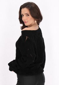 Maglione nero a maglia con lavorazione a rete aperta e design off-the-shoulder. Presenta polsini e orlo a coste, abbinato a pantaloni neri aderenti.