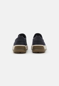 Marineblå treningssko med en glatt overflate, en hvit såle og en brun yttersåle. Har et slip-on design og minimale sømdetaljer.