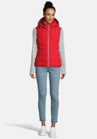 Vrouw in een rode doorgestikte mouwloze vest met capuchon over een wit en zwart gestreept langslavendshirt, met lichtblauwe jeans en witte sneakers.
