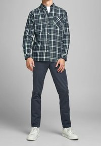 Groen- en donkerblauw geruit overhemd met knopen en een borstzak, gecombineerd met slim-fit donkere broek en witte sneakers. Effen achtergrond.