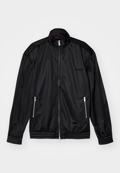 Svart glidelåsjakke i lett, glansete stoff. Har to glidelåslommer, stående krage og elastiske mansjetter og kant.