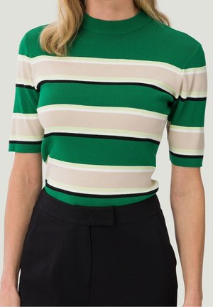 Femme portant un haut tricoté vert à manches courtes avec des rayures horizontales blanches, beiges et noires, rentrée dans un pantalon noir taille haute.