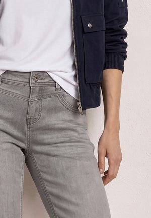 Osoba ubrana w jasnoszare dżinsy, białą koszulkę i granatową bluzę na zamek z kieszenią na guziki, ramieniem swobodnie opuszczonym wzdłuż ciała.