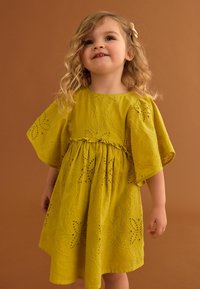 Gelbes, besticktes Kleid mit kurzen, glockenförmigen Ärmeln, das mit floralen Ausschnitten und einem gerafften Bund ausgestattet ist. Weicher Stoff vor einem braunen Hintergrund.