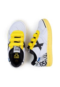 Zapatillas infantiles blancas y amarillas con detalles en negro, tres correas de Velcro amarillas y estampados gráficos en el talón y la suela.