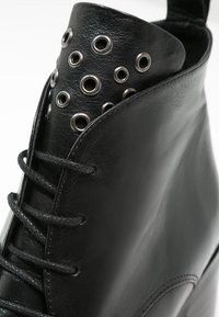 Botín de cuero negro hasta el tobillo con ojales de metal en el puño, cordones y una punta redondeada elegante. Textura suave con detalles de costura mínimos.