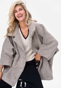 Vlasgrijze oversized jas met brede revers, gestructureerde stof en een knoopsluiting aan de voorkant. Draagt over een crèmekleurige V-hals trui en zwarte broek.
