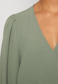 Top verde a V realizzato in tessuto testurizzato, con dettagli arricciati sulle spalle. Le maniche sono lunghe e leggermente aderenti.