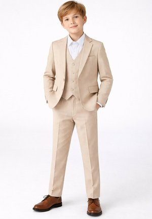 Garçon aux cheveux châtain clair portant un costume trois pièces beige, chemise blanche et chaussures de ville marron, debout les mains dans les poches.