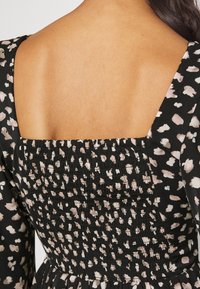 Górna część pleców kobiety, ukazująca skórę i czarną sukienkę z jasnoróżowymi i szarymi abstrakcyjnymi plamami, kwadratowym dekoltem i marszczonym detalem.