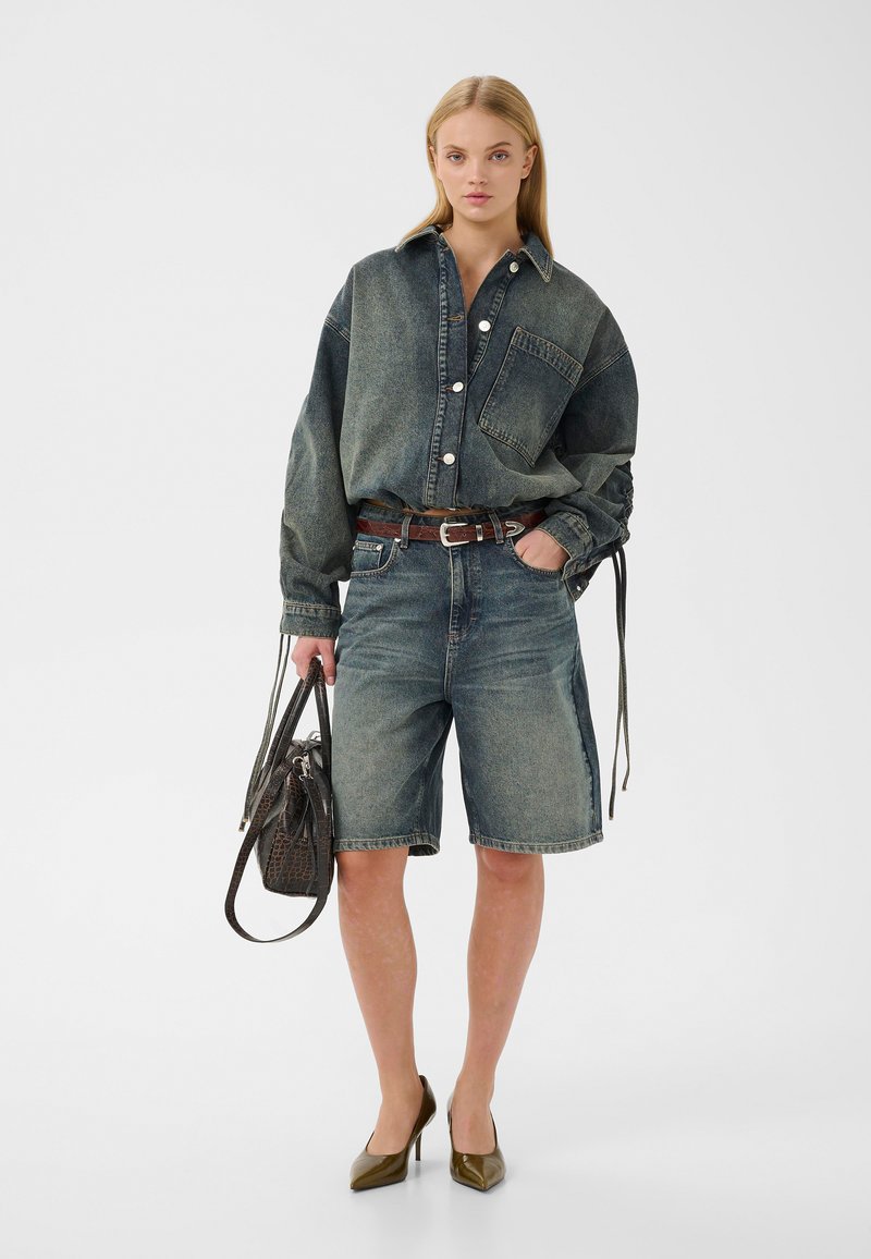 Femme portant une chemise en jean oversize lavée et un short long assorti, tenant un sac à main sombre et des talons hauts marron.