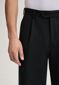 Pantalons de costume noirs au toucher lisse, dotés d'une fermeture à un bouton et de poches latérales, présentés avec une main reposant dessus.