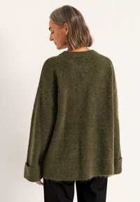 Vrouw met grijs schouderlang haar, gekleed in een oversized olijfgroen fuzzy sweater en zwarte broek, van achteren gezien.