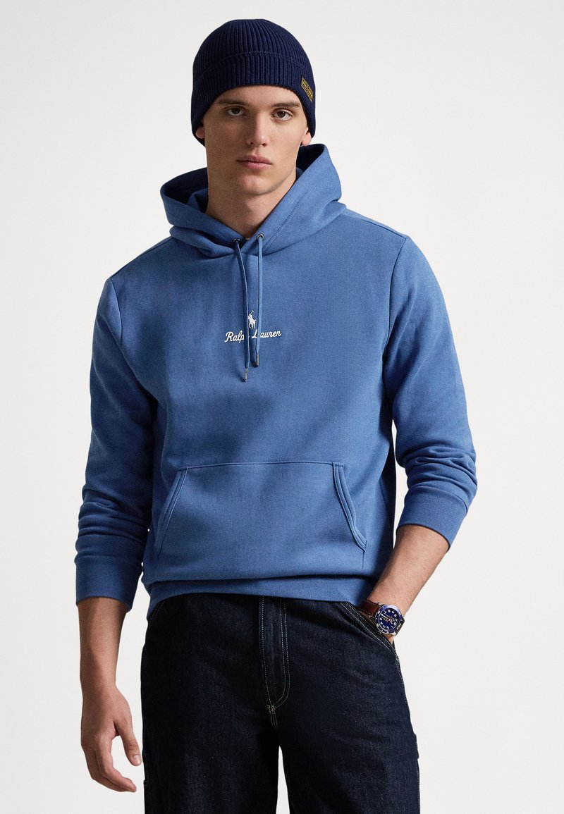 Blå bomullshoodie med känguruficka och dragsko i huvan. Har broderad logotyp och ribbade muddar; kombinerad med mörka jeans och en klocka.