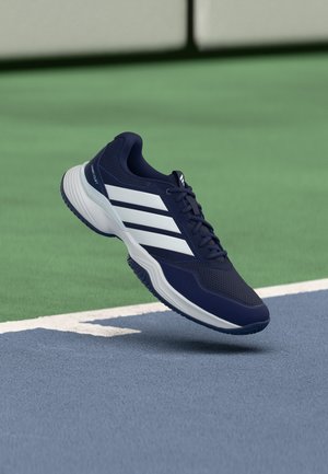 Scarpa sportiva blu navy con strisce bianche e suola bianca, posizionata in aria sopra la superficie di un campo da tennis.