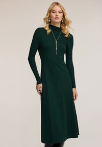 Abito lungo e aderente di colore verde scuro, con maniche lunghe a coste e scollatura alta. Tessuto morbido, gonna svasata. Collana con pendente argentato come dettaglio.