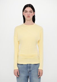 Gelbes Langarmoberteil mit Rundhalsausschnitt, das eine weiche Textur und geraffte Details an der taille aufweist, kombiniert mit hellblauen Jeans.