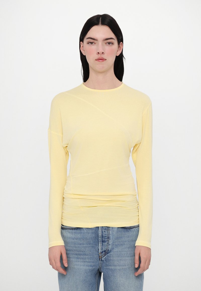 Gelbes Langarmoberteil mit Rundhalsausschnitt, das eine weiche Textur und geraffte Details an der taille aufweist, kombiniert mit hellblauen Jeans.