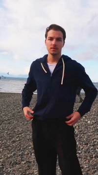 Granatowa bluza z zamkiem i kremowymi sznurkami, noszona na białej koszulce; czarne spodnie; stoi na kamienistej plaży blisko wody.