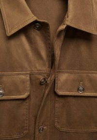 Chemise en daim marron avec col, deux poches avant et fermetures à boutons. Texture douce et détails de couture subtils qui mettent en valeur le design.