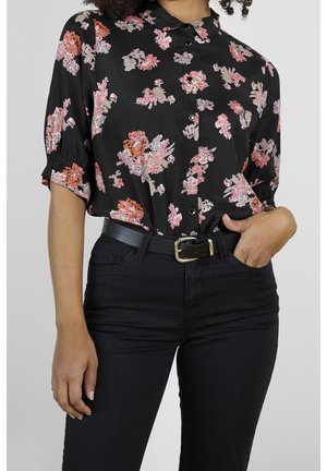 Zwart bloemenoverhemd met korte mouwen, een kraag en knoopsluiting. Draagt bij zwarte jeans en een zwarte riem met een gouden gesp.