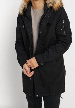 Mann trägt eine schwarze Parka mit einem flauschigen Kapuzenbesatz und durchgehendem Reißverschluss über einem beigen Pullover, steht vor einem einfarbigen Hintergrund.