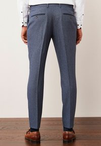 Blaue gemusterte Hosen mit schmaler Passform, zwei Gesäßtaschen mit Knöpfen und einer Bügelfalte an der Vorderseite. Braune Lederschuhe sind sichtbar.