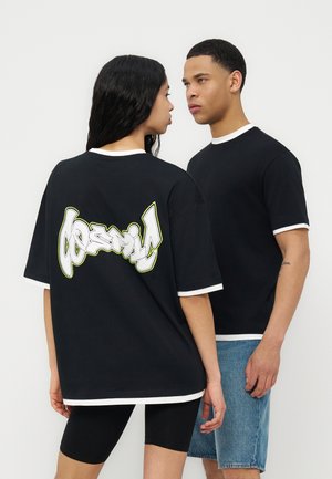 To personer iført sorte T-shirts med hvide kanter, hvoraf den ene viser en kraftig grafik på bagsiden, står og vender hinanden.