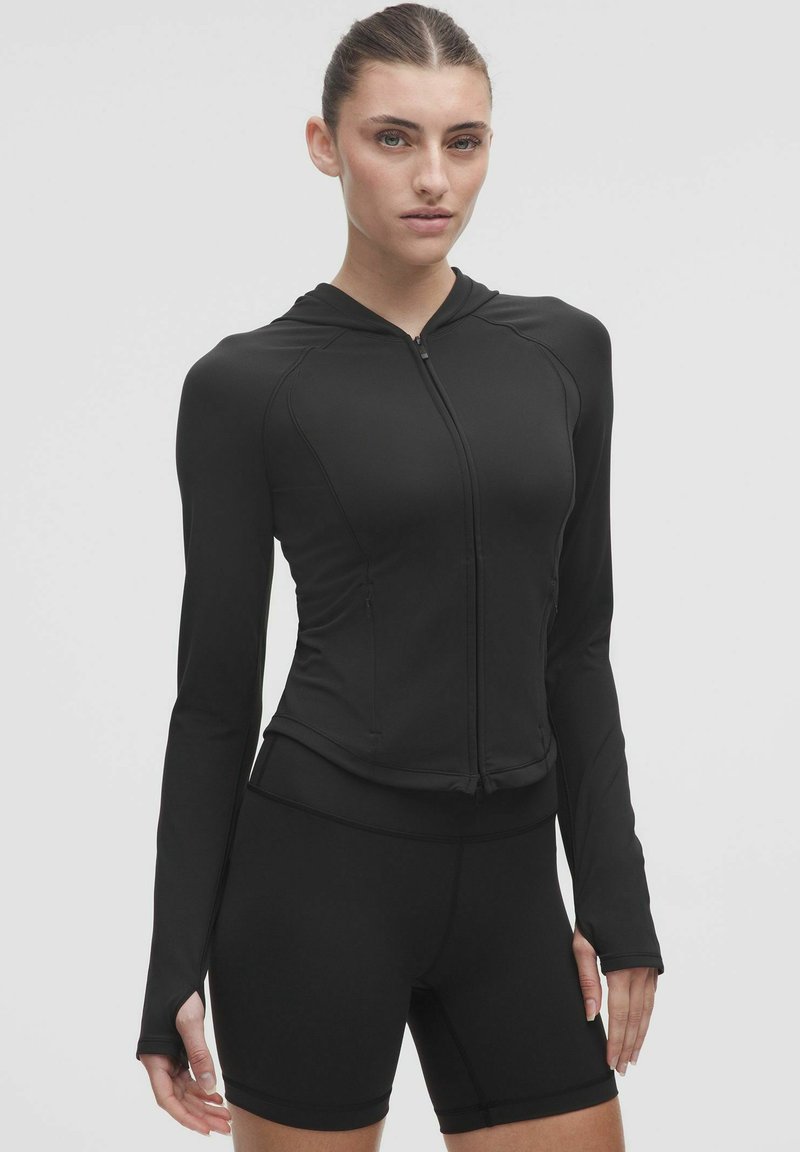 Jachetă sport neagră cu fermoar și glugă, mâneci lungi și un design adaptat. Fabricată dintr-un material neted și elastic, asociată cu pantaloni scurți negri asortați.