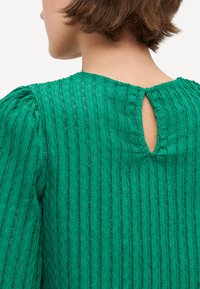 Green textured blouse with vertical stripes, featuring puffed sleeves and a keyhole opening at the back. Made from lightweight fabric.