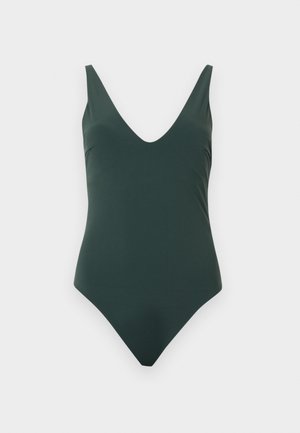 Tumeheline üheosaline ujumistrikoo V-kaelusega ja laiad õlapaelad ühtlase heleda tausta ees.