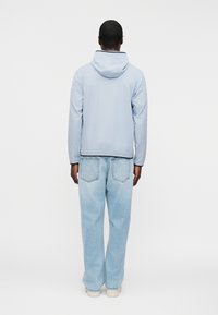 Άνδρας κοντά με γυρισμένη την πλάτη, φοράει ένα ανοιχτό μπλε μπουφάν με κουκούλα, άνετα τζιν ανοιχτού ξεβαμμένου χρώματος και λευκά αθλητικά παπούτσια, πάνω σε λευκό φόντο.