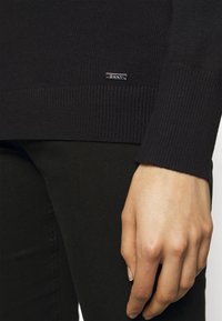 Svart stickad tröja med ribbade ärmar och nederkant, med en metalllogg på, bärs över svarta byxor. Slät textur, figurnära design.