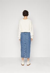 Sweatshirt blanc surdimensionné et jupe en denim bleu, longueur cheville, avec une fente au dos. Des chaussures blanches décontractées avec des semelles de couleur claire complètent la tenue.