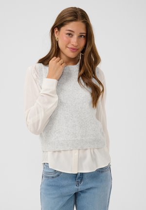 Joven mujer con cabello largo y castaño lleva blusa blanca, suéter sin mangas gris claro, vaqueros azules y pendientes de oro, sonriendo a la cámara.