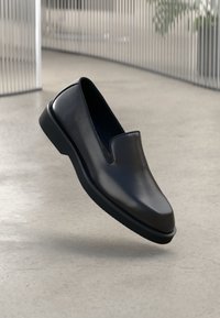 Sorte lædersslip-on sko med rund tå, forhøjet frontpanel og struktureret gummisål, placeret mod en glat gulv.