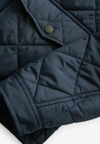 Marineblau gesteppte Jacke mit glatter Textur, ausgestattet mit einem Druckknopf-Akzent und geometrischem Muster aus Rautennähten auf dem Stoff.