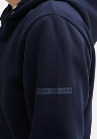Marineblauwe hoodie van een gestructureerde stof, met een logo patch op de mouw met "TOM TAILOR" en extra tekst eronder.